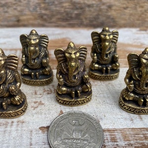 Op de afbeelding: Vijf kleine koperen beeldjes van de godheid Ganesha, elk zittend op een ronde voet met gedetailleerde versieringen. Een Amerikaanse kwartmunt is op de voorgrond geplaatst om een idee te geven van de schaal van de beeldjes.