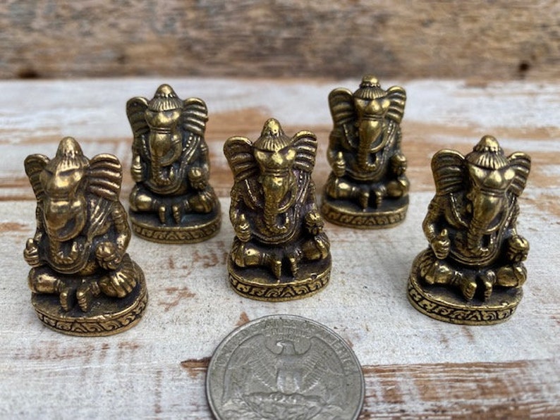 Op de afbeelding: Vijf kleine koperen beeldjes van de godheid Ganesha, elk zittend op een ronde voet met gedetailleerde versieringen. Een Amerikaanse kwartmunt is op de voorgrond geplaatst om een idee te geven van de schaal van de beeldjes.
