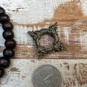 Pu&ograve; includere: Un anello in metallo color bronzo, ornato con decorazioni a forma di drago. L'anello &egrave; di forma quadrata con un'apertura circolare al centro. Una moneta &egrave; accanto all'anello per il confronto delle dimensioni. Perline di legno marrone scuro sono visibili a sinistra.