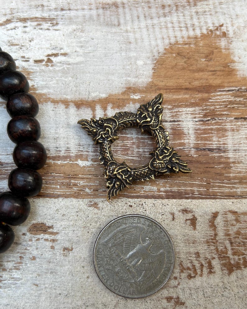 Pu&ograve; includere: Un anello in metallo color bronzo, ornato con decorazioni a forma di drago. L'anello &egrave; di forma quadrata con un'apertura circolare al centro. Una moneta &egrave; accanto all'anello per il confronto delle dimensioni. Perline di legno marrone scuro sono visibili a sinistra.