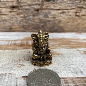 Op de afbeelding: Een klein, goudkleurig messing beeldje van de godheid Ganesha, zittend afgebeeld met een olifantenkop en grote oren. Het beeldje staat op een ronde voet met decoratieve gravures en is naast een muntstuk geplaatst.