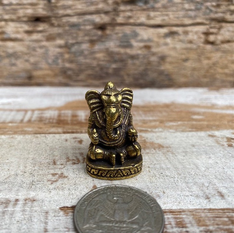Op de afbeelding: Een klein, goudkleurig messing beeldje van de godheid Ganesha, zittend afgebeeld met een olifantenkop en grote oren. Het beeldje staat op een ronde voet met decoratieve gravures en is naast een muntstuk geplaatst.