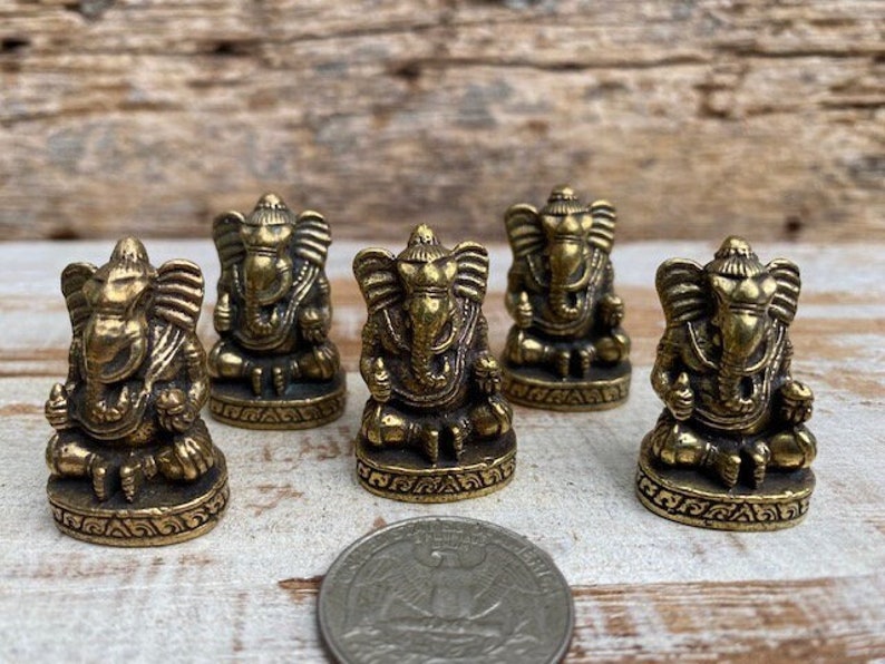 Op de afbeelding: Vijf kleine koperen Ganesha-beeldjes in zittende positie, elk met een olifantenkop, grote oren en gedetailleerde versieringen op de basis. Een Amerikaanse kwartje ligt op de voorgrond om de schaal van de beeldjes aan te geven.