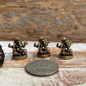K&ouml;nnte beinhalten: Drei kleine, antik goldfarbene Ganesh-Statuen sind auf einer Holzoberfl&auml;che angeordnet. Eine M&uuml;nze befindet sich neben den Statuen zum Gr&ouml;&szlig;envergleich. Die Statuen sind detailliert mit aufwendigen Designs und stellen die hinduistische Gottheit dar.
