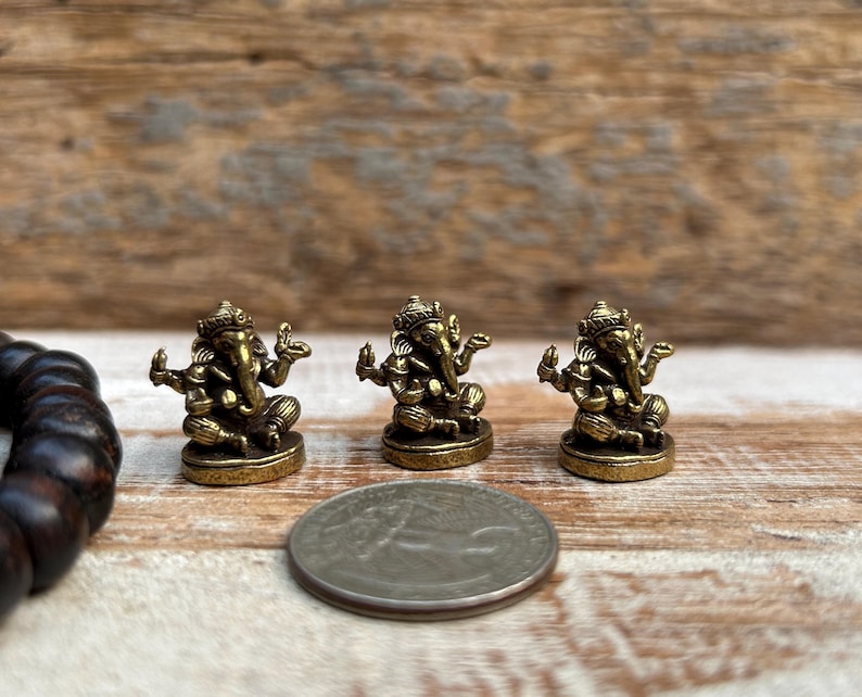 K&ouml;nnte beinhalten: Drei kleine, antik goldfarbene Ganesh-Statuen sind auf einer Holzoberfl&auml;che angeordnet. Eine M&uuml;nze befindet sich neben den Statuen zum Gr&ouml;&szlig;envergleich. Die Statuen sind detailliert mit aufwendigen Designs und stellen die hinduistische Gottheit dar.