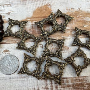 Pu&ograve; includere: Una collezione di cornici decorative quadrate color ottone antico con intricati disegni di draghi e fiori. Le cornici sono disposte su una superficie di legno chiaro, con una collana di perline di legno scuro e una moneta.
