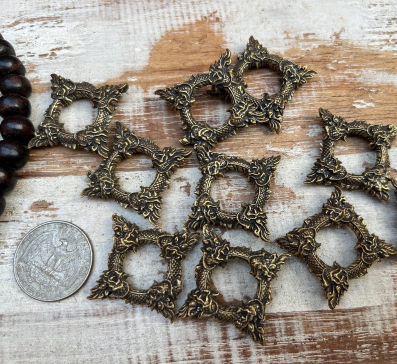 Pu&ograve; includere: Una collezione di cornici decorative quadrate color ottone antico con intricati disegni di draghi e fiori. Le cornici sono disposte su una superficie di legno chiaro, con una collana di perline di legno scuro e una moneta.