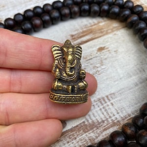 Op de afbeelding: Een klein, goudkleurig metalen beeldje van de godheid Ganesha, afgebeeld in een zittende houding met een olifantenkop en grote oren op een decoratieve voet, vergezeld door een lange streng donkerbruine, bolvormige houten gebedskralen.