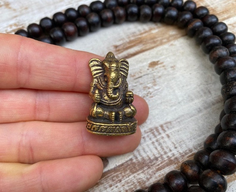 Op de afbeelding: Een klein, goudkleurig metalen beeldje van de godheid Ganesha, afgebeeld in een zittende houding met een olifantenkop en grote oren op een decoratieve voet, vergezeld door een lange streng donkerbruine, bolvormige houten gebedskralen.