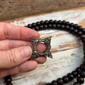 Pu&ograve; includere: Un ciondolo quadrato in ottone antico con un design a drago. Il ciondolo ha un'apertura circolare centrale ed &egrave; tenuto in mano. Una collana di perline marrone scuro &egrave; sullo sfondo.