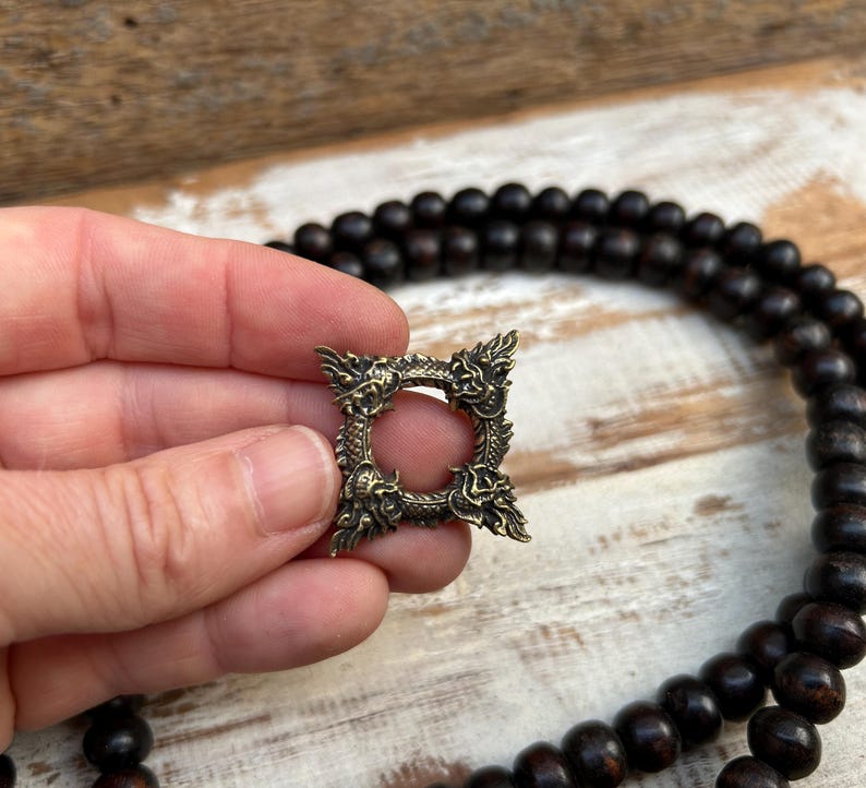 Pu&ograve; includere: Un ciondolo quadrato in ottone antico con un design a drago. Il ciondolo ha un'apertura circolare centrale ed &egrave; tenuto in mano. Una collana di perline marrone scuro &egrave; sullo sfondo.