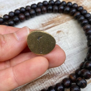Op de afbeelding: Een kleine, onregelmatige, goudkleurige metalen schijf met een geborstelde afwerking en een donkere vlek in het midden, naast een lange streng donkerbruine, bolvormige houten kralen in een dubbele lus op een lichtgekleurde houten ondergrond.