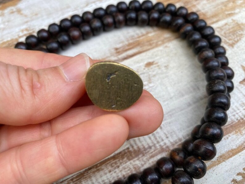 Op de afbeelding: Een kleine, onregelmatige, goudkleurige metalen schijf met een geborstelde afwerking en een donkere vlek in het midden, naast een lange streng donkerbruine, bolvormige houten kralen in een dubbele lus op een lichtgekleurde houten ondergrond.