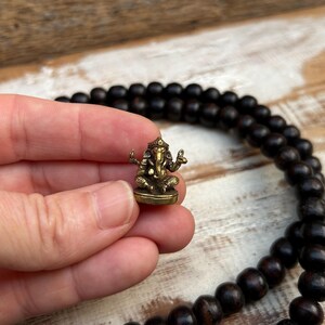 K&ouml;nnte beinhalten: Eine kleine Ganesh-Statue aus Messing wird in einer Hand gehalten, mit einer dunklen Holzperlenkette im Hintergrund. Die Statue zeigt die hinduistische Gottheit Ganesh in sitzender Position mit mehreren Armen und einem Elefantenkopf.