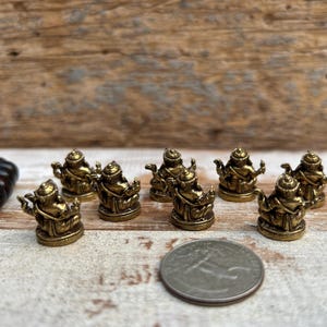 K&ouml;nnte beinhalten: Eine Sammlung kleiner, goldfarbener Buddha-Statuen. Die Statuen sind auf einer hellen Oberfl&auml;che mit einem Holz Hintergrund angeordnet. Eine M&uuml;nze ist als Gr&ouml;&szlig;enreferenz enthalten. Die Statuen sind detailliert und scheinen aus Metall zu sein.