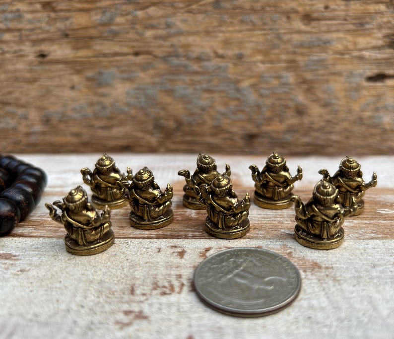 K&ouml;nnte beinhalten: Eine Sammlung kleiner, goldfarbener Buddha-Statuen. Die Statuen sind auf einer hellen Oberfl&auml;che mit einem Holz Hintergrund angeordnet. Eine M&uuml;nze ist als Gr&ouml;&szlig;enreferenz enthalten. Die Statuen sind detailliert und scheinen aus Metall zu sein.