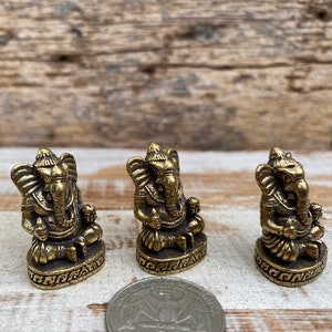 Op de afbeelding: Drie kleine messingkleurige metalen beeldjes van de godheid Ganesha, elk zittend op een ronde voet met decoratieve gravures. Een Amerikaanse kwartmunt is op de voorgrond geplaatst als schaalvergroting voor de drie kleine beelden.