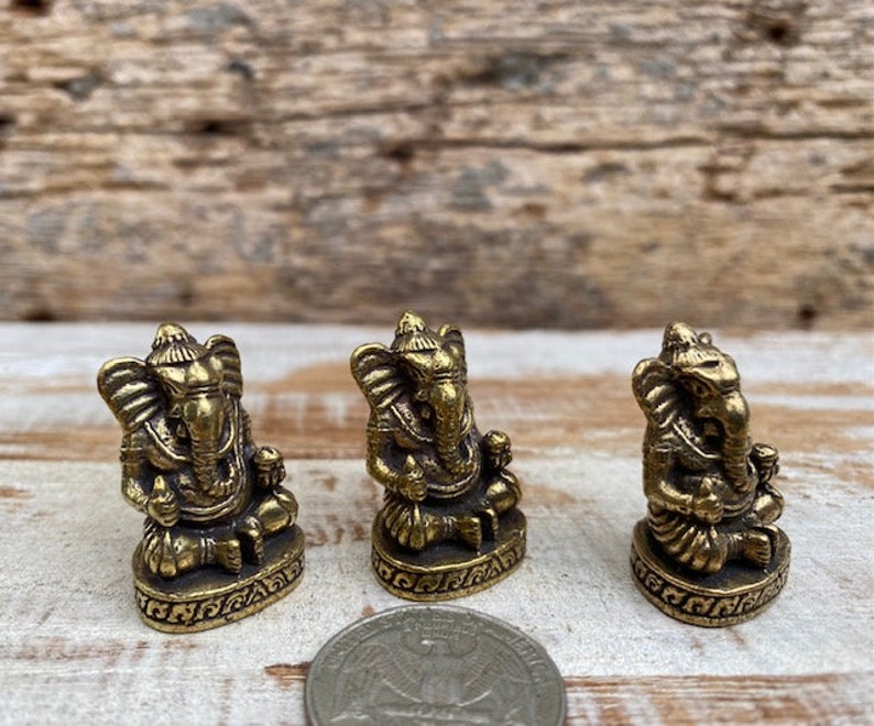 Op de afbeelding: Drie kleine messingkleurige metalen beeldjes van de godheid Ganesha, elk zittend op een ronde voet met decoratieve gravures. Een Amerikaanse kwartmunt is op de voorgrond geplaatst als schaalvergroting voor de drie kleine beelden.