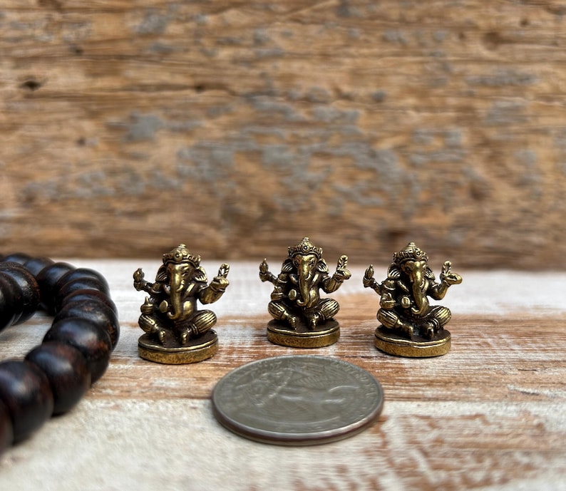 K&ouml;nnte beinhalten: Drei kleine, goldfarbene Ganesh-Statuen werden auf einer Holzoberfl&auml;che ausgestellt. Die Statuen sind aufwendig detailliert und sitzen in einer sitzenden Position. Ein dunkelbraunes Perlenarmband und eine M&uuml;nze sind ebenfalls im Bild.