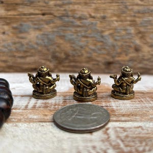 K&ouml;nnte beinhalten: Drei kleine, goldfarbene Statuen einer sitzenden Gottheit mit mehreren Armen und einem detaillierten Kopfschmuck. Eine M&uuml;nze liegt neben den Statuen zum Gr&ouml;&szlig;envergleich. Ein dunkelbraunes Perlenarmband befindet sich im Hintergrund. Die Statuen stehen auf einer Holzoberfl&auml;che.