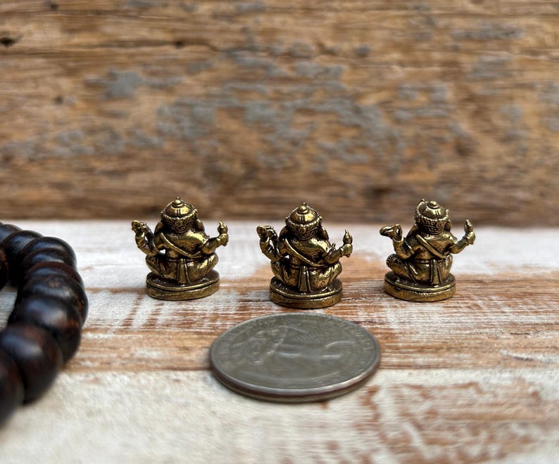 K&ouml;nnte beinhalten: Drei kleine, goldfarbene Statuen einer sitzenden Gottheit mit mehreren Armen und einem detaillierten Kopfschmuck. Eine M&uuml;nze liegt neben den Statuen zum Gr&ouml;&szlig;envergleich. Ein dunkelbraunes Perlenarmband befindet sich im Hintergrund. Die Statuen stehen auf einer Holzoberfl&auml;che.