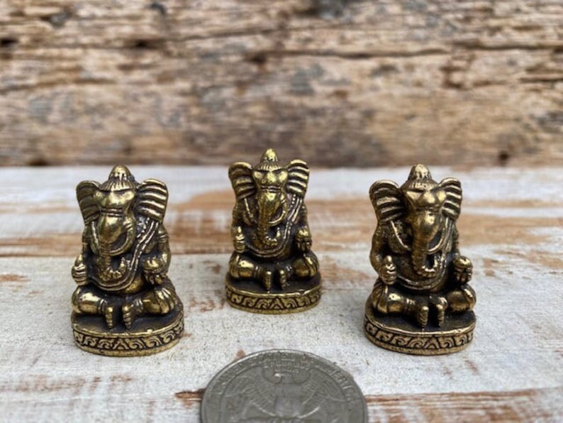 Op de afbeelding: Drie kleine, antiek ogende messing beeldjes van de godheid Ganesha, elk zittend afgebeeld met een olifantenkop en grote oren. De beeldjes staan in een rij opgesteld, met een muntstuk op de voorgrond voor een indicatie van de schaal.