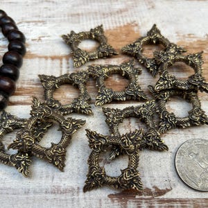 Pu&ograve; includere: Una collezione di cornici quadrate antiche color ottone con elaborati disegni a forma di drago. Le cornici sono sparse su una superficie di legno chiaro, insieme a una collana di perline di legno scuro e una moneta d'argento.