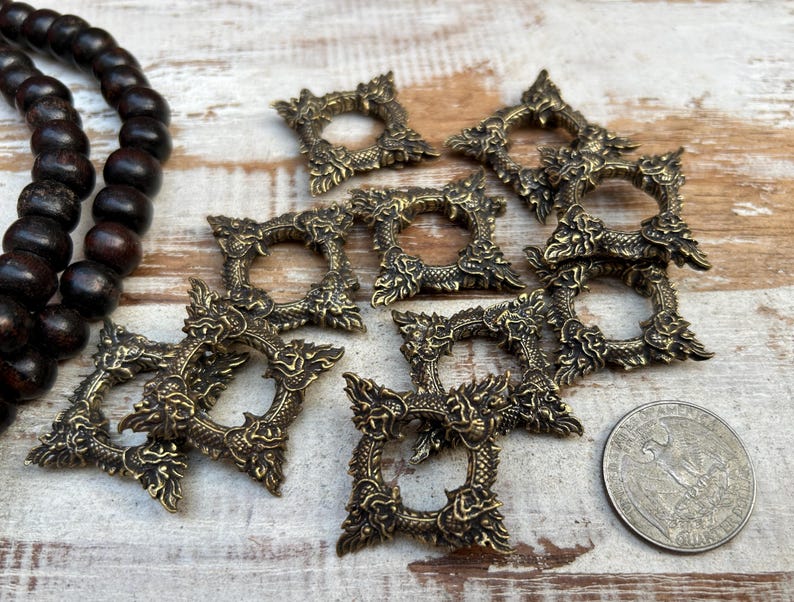 Pu&ograve; includere: Una collezione di cornici quadrate antiche color ottone con elaborati disegni a forma di drago. Le cornici sono sparse su una superficie di legno chiaro, insieme a una collana di perline di legno scuro e una moneta d'argento.