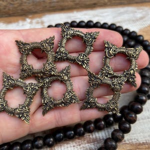 Pu&ograve; includere: Cinque anelli decorativi in metallo color ottone antico con disegni di draghi. Gli anelli sono quadrati con un'apertura circolare al centro. I dettagli del drago sono intricati e ornati. Una collana di perline di legno scuro &egrave; sullo sfondo.