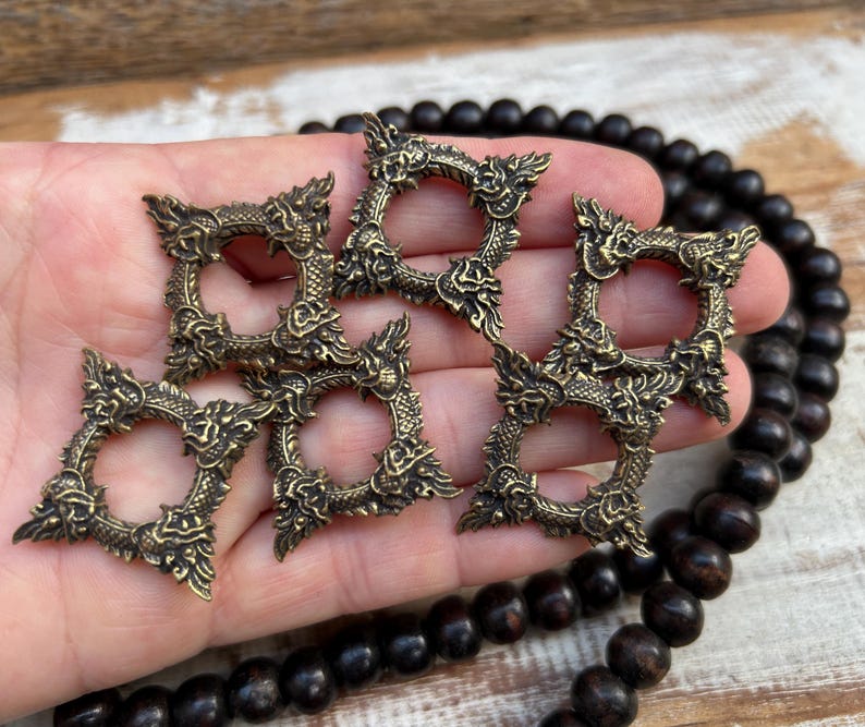 Pu&ograve; includere: Cinque anelli decorativi in metallo color ottone antico con disegni di draghi. Gli anelli sono quadrati con un'apertura circolare al centro. I dettagli del drago sono intricati e ornati. Una collana di perline di legno scuro &egrave; sullo sfondo.
