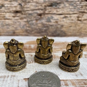 Op de afbeelding: Drie kleine messingkleurige beeldjes van een zittende godheid met olifantachtige kenmerken, van achteren gezien. Elk figuur rust op een ronde basis met decoratieve gravures. Een zilverkleurige Amerikaanse kwartmunt ligt op de voorgrond.