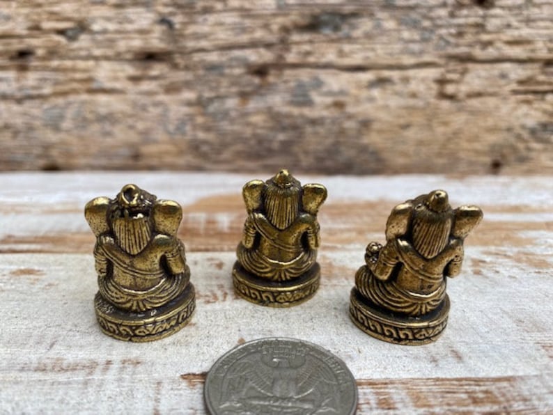 Op de afbeelding: Drie kleine messingkleurige beeldjes van een zittende godheid met olifantachtige kenmerken, van achteren gezien. Elk figuur rust op een ronde basis met decoratieve gravures. Een zilverkleurige Amerikaanse kwartmunt ligt op de voorgrond.