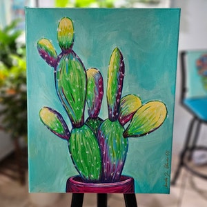 Könnte beinhalten: Ein lebendiges Gemälde eines Kaktus in einem roten Topf vor türkisem Hintergrund. Der Kaktus hat grüne, gelbe und violette Farbtöne mit weißen Sprenkeln. Das Kunstwerk wird auf einer schwarzen Staffelei präsentiert, die ein botanisches Thema zeigt.