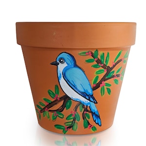 May include: A terracotta flower pot with a hand-painted bluebird perched on a branch with green leaves and red berries. The bird has blue and white feathers. The pot is a classic, tapered shape.