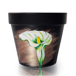Könnte beinhalten: Schwarz bemalter Blumentopf mit einem handgemalten Design einer weißen Calla. Die Blume hat eine gelbe Mitte und grüne Akzente. Der Hintergrund hat ein bronzefarbenes Pinselstrich-Design.