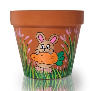 Puede incluir: Una maceta de terracota con un diseño pintado a mano de un conejo marrón sosteniendo una zanahoria. El conejo tiene orejas rosas y hierba verde lo rodea.