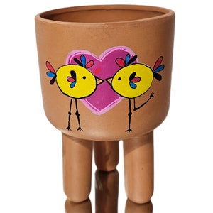 May include: A terracotta planter with three legs features a hand-painted design of two yellow birds kissing in front of a pink heart.