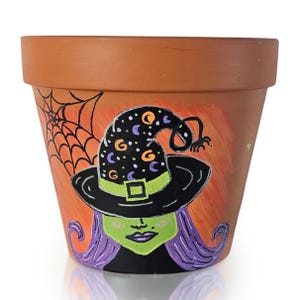 Peut inclure: Un pot de fleur en terre cuite avec un motif de sorcière d'Halloween peint à la main. La sorcière a la peau verte, des cheveux violets et un chapeau noir orné de lunes et d'étoiles. Une toile d'araignée est peinte sur le côté, sur fond orange.