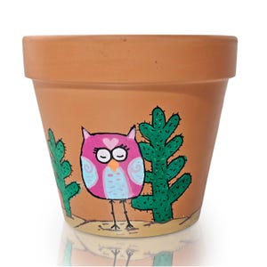 May include: A terracotta-colored flower pot with a hand-painted design. The design features a pink owl with a heart on its chest, blue accents, and closed eyes, flanked by green cacti. The pot is suitable for small plants.