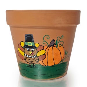 May include: A terracotta-colored flower pot with a hand-painted Thanksgiving design. The design features a cartoon turkey wearing a pilgrim hat next to an orange pumpkin. The base of the pot has a green painted border.