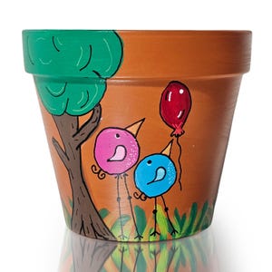 Puede incluir: Una maceta de terracota pintada con un árbol verde, dos pájaros y un globo rojo. Los pájaros son rosa y azul y tienen patas largas. La maceta está decorada con hojas verdes.