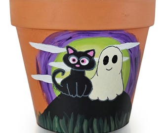 以下が含まれることがあります： テラコッタ色の植木鉢。手描きのハロウィーンのデザインで、ピンクの目をした黒猫と白い幽霊が、紫と緑の背景に描かれています。小さな植物や季節の装飾に適しています。