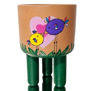May include: A terracotta planter with three legs, painted with a pink heart and two birds. The larger bird is purple with a yellow beak and the smaller bird is yellow with a pink beak. The planter is a great addition to any home decor.