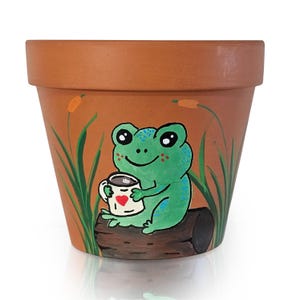 May include: A terracotta-colored flower pot with a hand-painted design. A green frog with blue spots sits on a log, holding a mug with a heart. The pot features cattails and grass painted on the side.