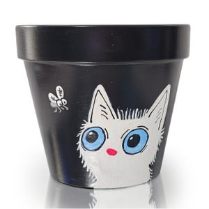 Puede incluir: Maceta de cerámica pintada de negro con un diseño de gato caprichoso. El gato tiene pelaje blanco, ojos azules grandes y una nariz roja. Una pequeña mosca está pintada en el lateral. Adecuada para uso en interiores o exteriores.