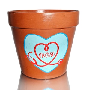 Puede incluir: Maceta de terracota con un corazón azul y un diseño de estetoscopio rojo. La palabra "Nurse" está escrita dentro del corazón.