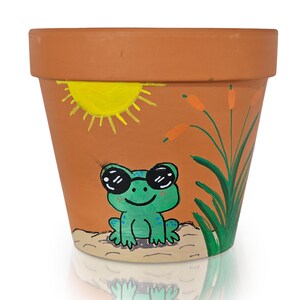 以下が含まれることがあります： 手描きのデザインが施されたテラコッタ製の植木鉢。サングラスをかけた笑顔の緑色のカエル、黄色の太陽、ガマが描かれています。カエルは砂地の上に座っています。この鉢は、花やハーブを植えるのに適しています。