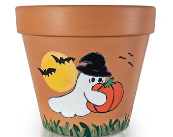以下が含まれることがあります： テラコッタ色の植木鉢。ハロウィーンをテーマにしたデザインで、魔女の帽子をかぶった漫画の幽霊がオレンジ色のカボチャを持っています。コウモリのいる黄色い月と緑の草がデザインを完成させています。
