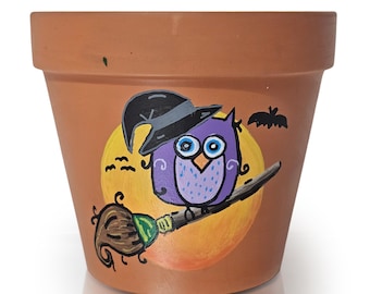 以下が含まれることがあります： ハロウィーンをテーマにしたデザインで装飾されたテラコッタ色の植木鉢。デザインは、魔女の帽子をかぶった紫色のフクロウが、黄色の月の前でほうきに乗っている様子を描いています。黒いコウモリと小さな鳥もポットに描かれています。