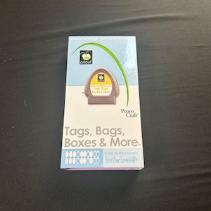 Könnte beinhalten: Eine rechteckige Cricut Formen-Kartuschenschachtel mit dem Titel Tags, Bags, Boxes & More. Die Verpackung ist weiß und hellblau mit einer grünen geschwungenen Grafik, dem Provo Craft-Logo und dem Namen Heather Lancaster in der rechten Ecke.